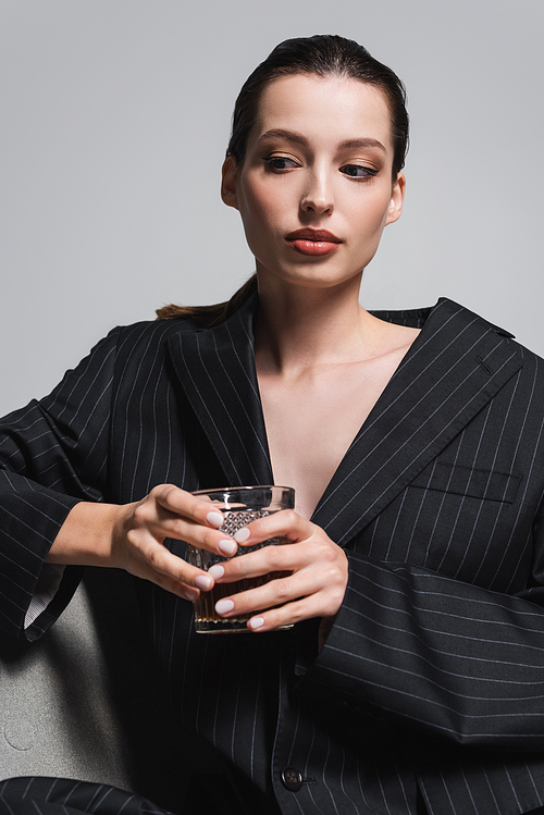 Portrait of woman in striped jacket holding glass of whiskey while sitting on armchair isolated on grey