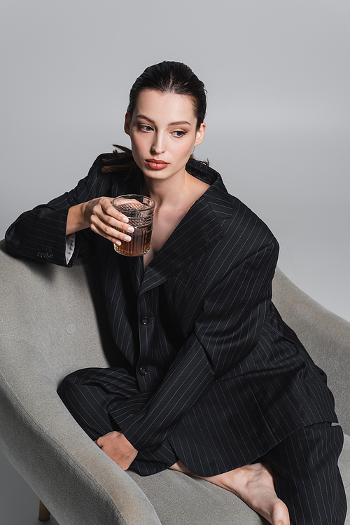 Brunette woman in striped suit holding whiskey while sitting on armchair on grey background