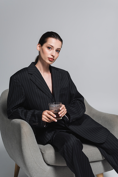 Stylish woman in striped suit holding whiskey and looking at camera on armchair on grey background