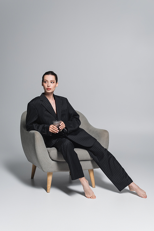Barefoot woman in black suit holding whiskey glass on armchair on grey background