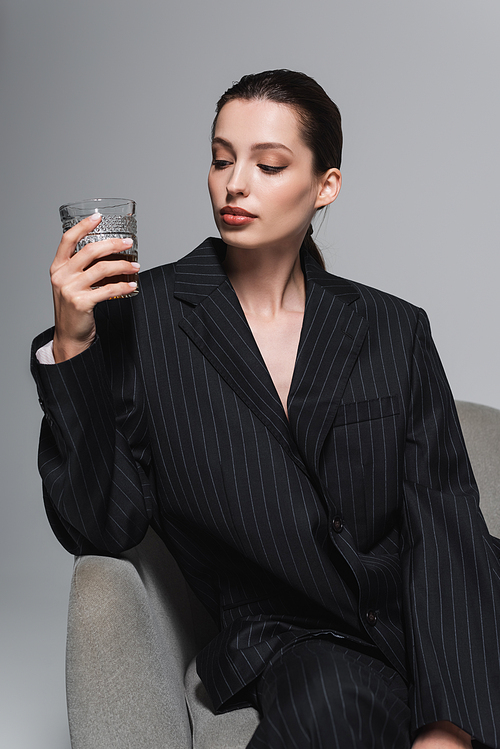Stylish woman in striped jacket looking at glass of whiskey on armchair isolated on grey