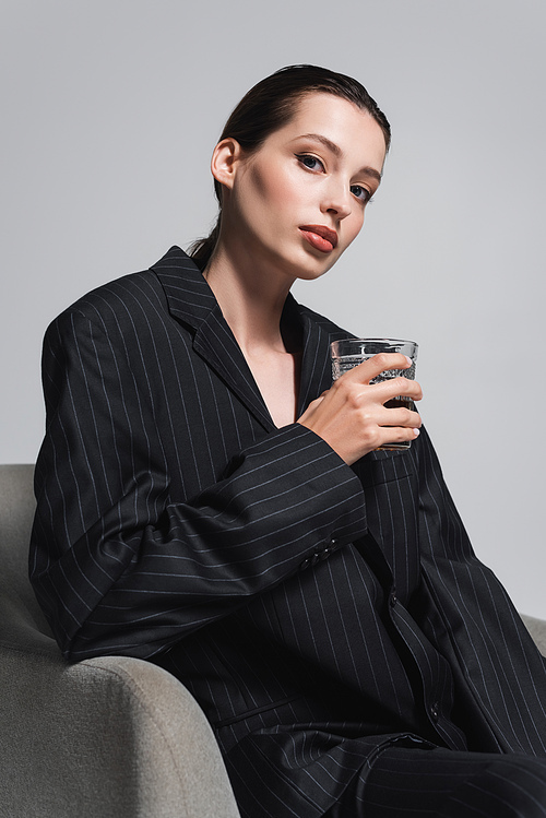 Brunette model in stylish suit holding glass of bourbon while sitting on armchair isolated on grey