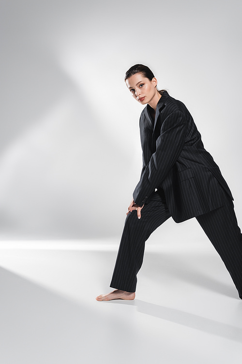 Trendy barefoot woman in suit posing and looking at camera on abstract grey background