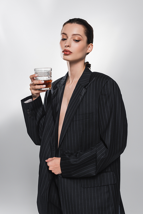 Stylish young woman in striped jacket holding whiskey on abstract grey background