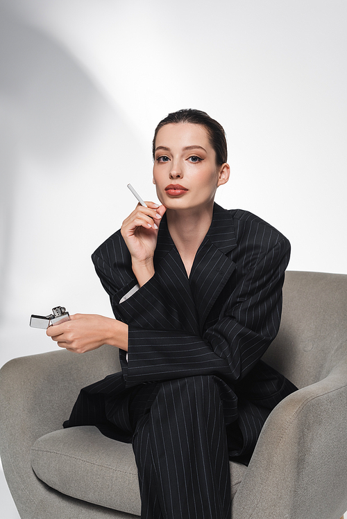 Trendy woman in striped suit holding cigarette and lighter while sitting on armchair on abstract grey background