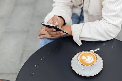 partial view of african american man texting on smartphone near cup of cappuccino on table
