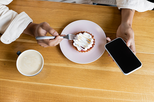 top view of african american man holding smartphone near tasty tart and cup of cappuccino