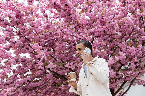 young african american man holding takeaway drink and talking on smartphone near pink cherry tree