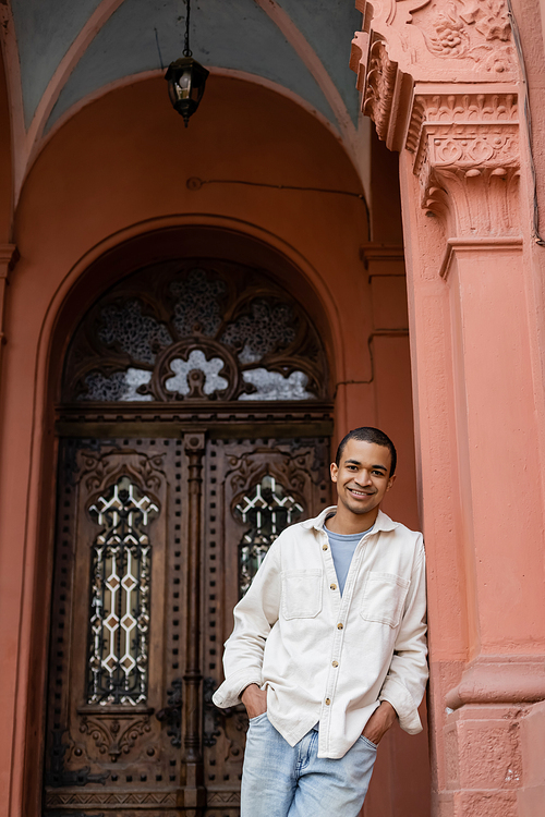 happy african american man in shirt jacket posing with hands in pockets near building in europe