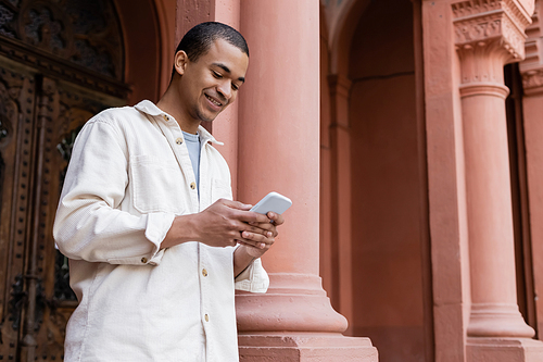 cheerful african american man in stylish shirt jacket messaging on smartphone near building