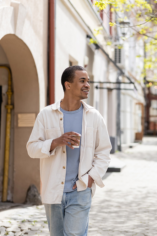 happy african american man in shirt jacket holding takeaway drink and walking with hand in pocket in urban city