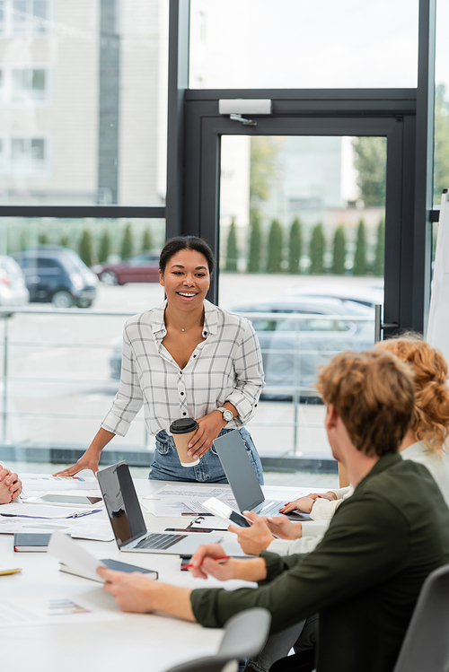 happy multiracial woman with paper cup talking to business team during meeting