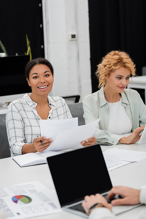 brunette multiracial businesswoman holding documents near blonde colleague and laptop with blank screen on blurred foreground