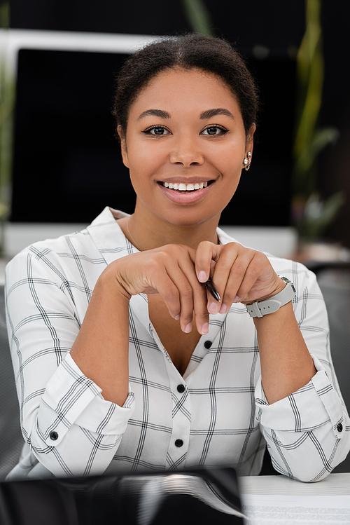 happy multiracial businesswoman holding pen and smiling at camera in office