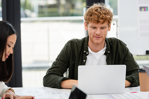 young redhead businessman working at laptop near asian colleague