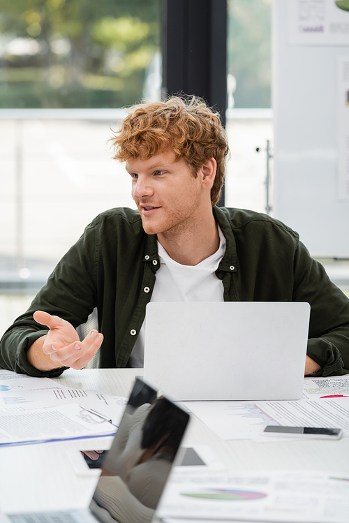 redhead businessman pointing with hand and talking near laptops in office
