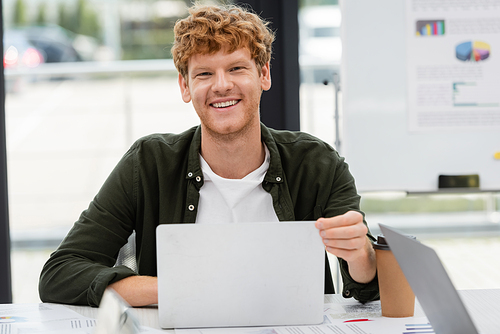 cheerful redhead manager smiling at camera near laptop and paper cup at workplace