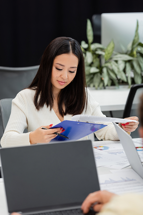 young asian businesswoman looking at documents on clipboard while working in office