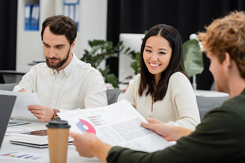 cheerful asian businesswoman looking at camera near colleagues working with documents in office