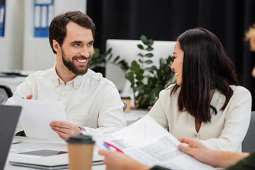 bearded man with document looking at young asian colleague while working in office