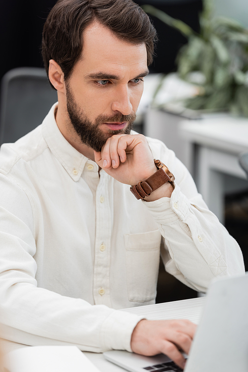 thoughtful bearded businessman holding hand near face and looking at blurred laptop