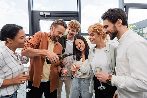 bearded man pouring champagne during corporate party with cheerful multiethnic colleagues