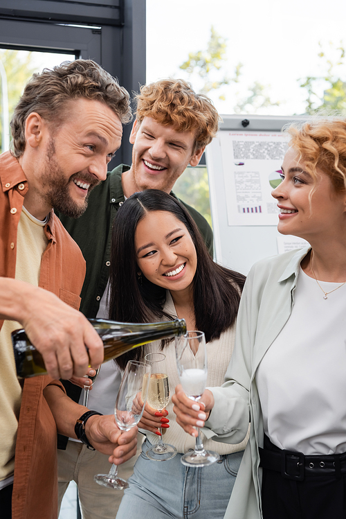 excited man pouring champagne near happy multicultural business colleagues during corporate party