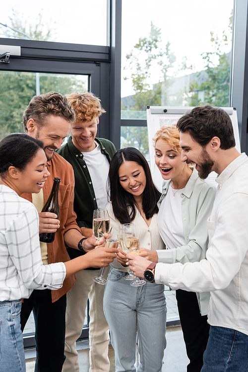 Happy multiethnic business people holding glasses near colleague with bottle of champagne in office
