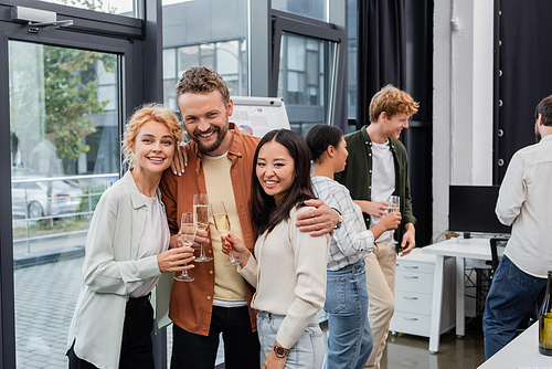 Positive businessman hugging multiethnic colleagues with champagne in office