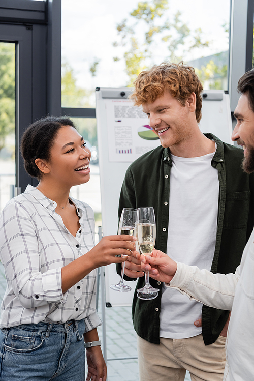 Cheerful multiracial businesswoman clinking champagne with colleagues in office