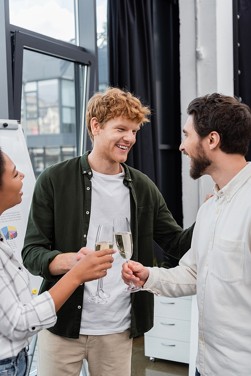 Smiling redhead businessman talking to interracial colleagues with champagne in office