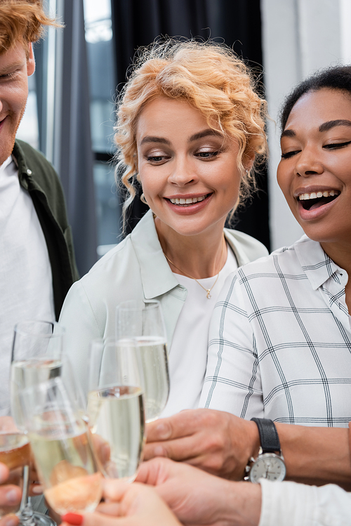 Positive businesswoman clinking champagne with interracial colleagues during corporate party in office