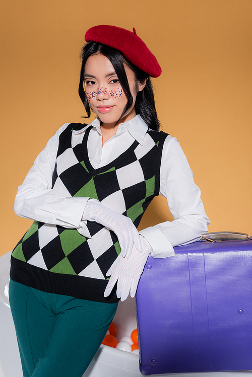 Trendy asian woman in gloves and beret standing near retro suitcase and bathtub with balls isolated on orange