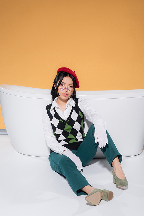 Fashionable asian woman in beret and gloves sitting near bathtub on orange background