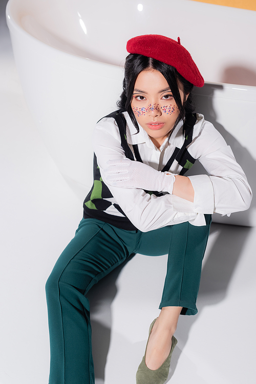 High angle view of pretty asian woman in beret looking at camera near bathtub on orange background
