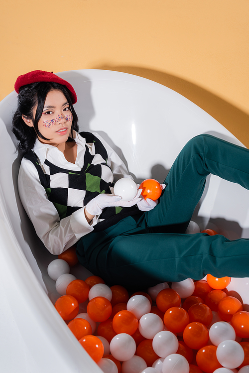 High angle view of stylish asian woman holding balls while sitting in bathtub on orange background