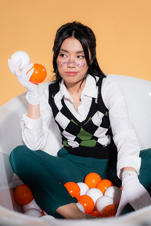 Stylish asian woman in gloves holding balls in bathtub isolated on orange