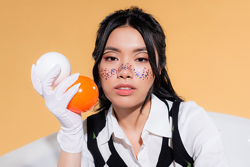 Portrait of young asian model with glitter on face holding balls and looking at camera in bathtub isolated on orange