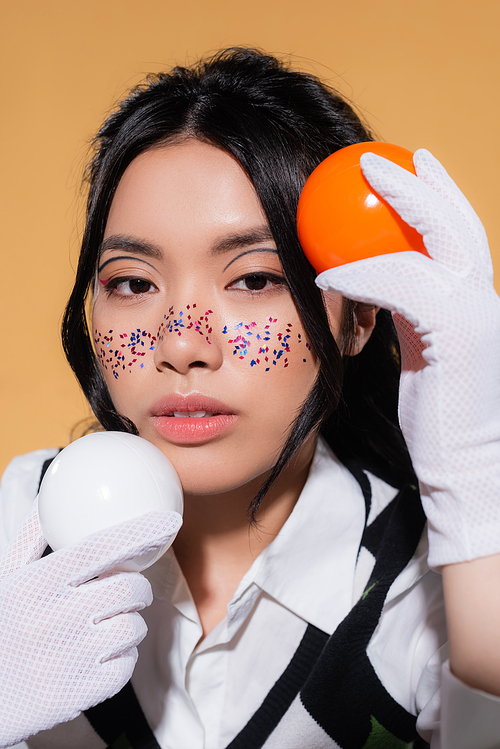 Portrait of asian model with glitter visage holding balls and looking at camera isolated on orange