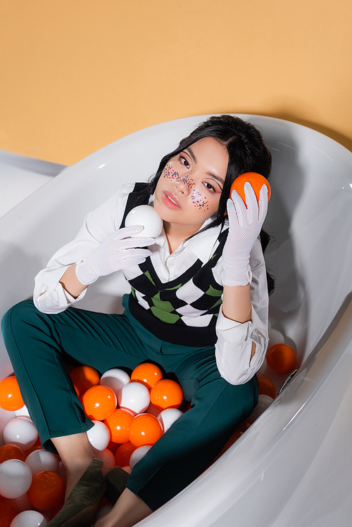 High angle view of trendy asian model in gloves holding balls while sitting in bathtub on orange background