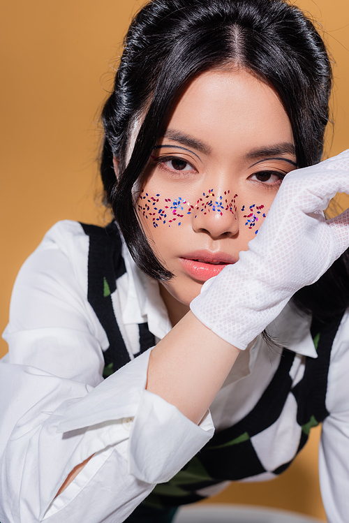 Portrait of fashionable asian model with glitter makeup looking at camera on orange background