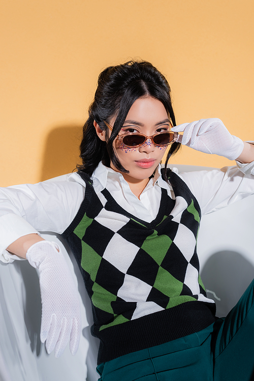 Asian model in sunglasses and vintage clothes sitting in bathtub on orange background