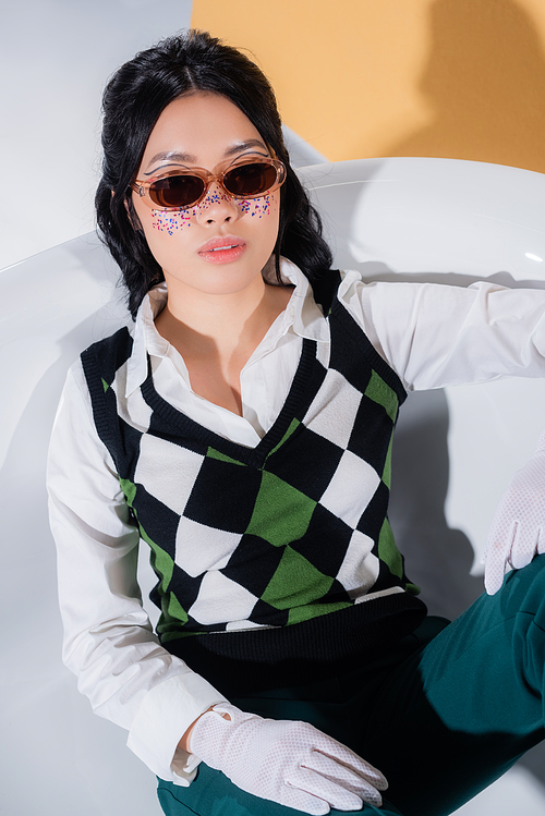 Asian model in sunglasses and retro clothes sitting in bathtub on orange background