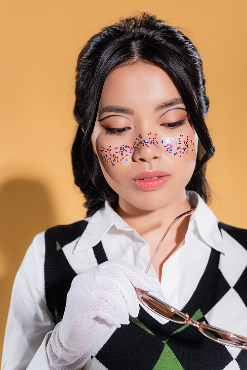 Young asian model with glitter on face holding sunglasses on orange background