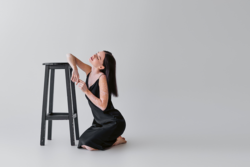 Sensual woman with vitiligo posing near chair on grey background