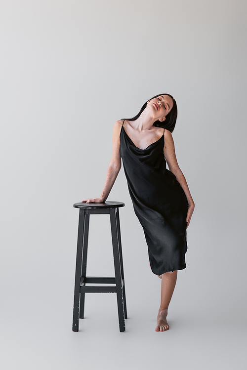 Sensual woman with vitiligo in silk dress touching chair on grey background