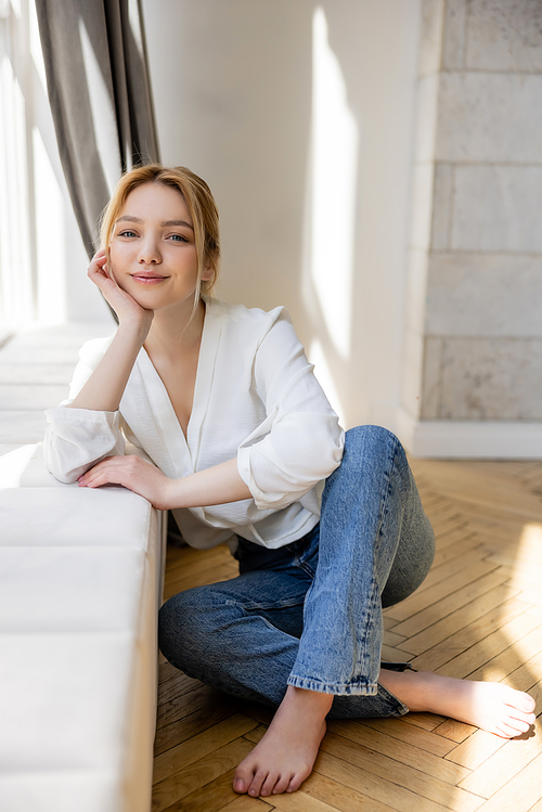 Barefoot woman in jeans smiling at camera near windowsill at home