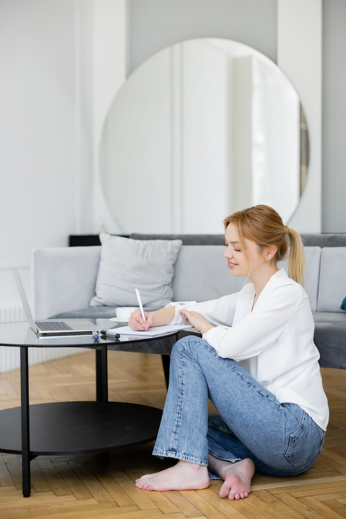 Cheerful barefoot freelancer writing on notebook near laptop in living room