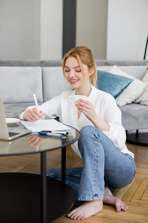 Pretty blonde freelancer holding cup and writing on notebook near laptop on coffee table in living room