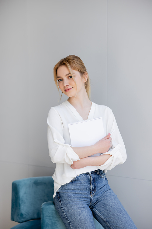 Blonde woman in blouse and jeans holding notebook near armchair at home